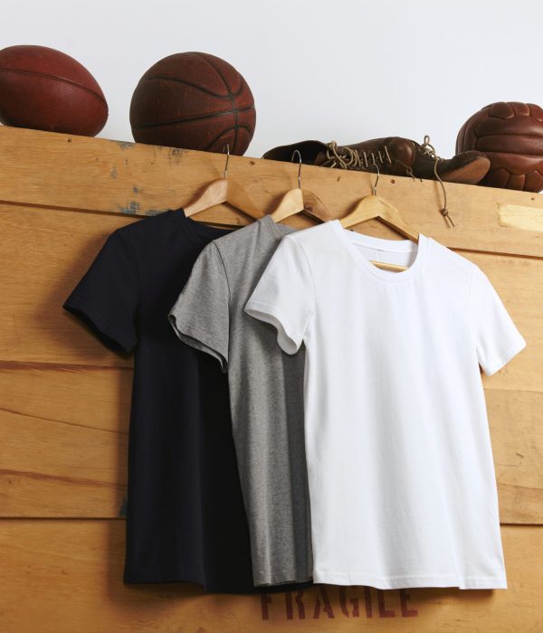 Presentation of a white, gray and black plain shortsleeve t-shirts with vintage football, basketball and volleyball and old sports leather boots on top of a wooden shipping box